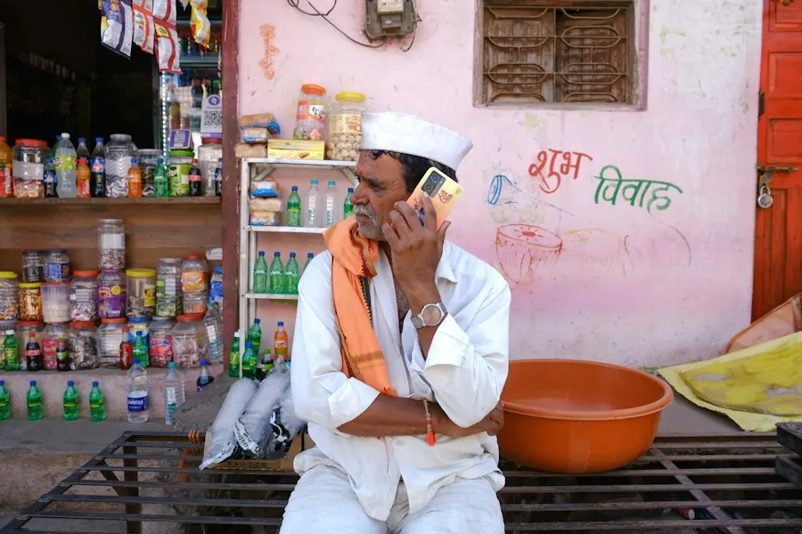 A shopkeeper in a white cap on a phone call outside his shop.